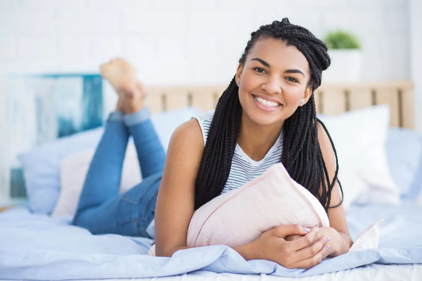 Smiling woman with braided hair lying on a bed, holding a pillow, in a bright and cozy bedroom setting, emphasizing comfort and relaxation.