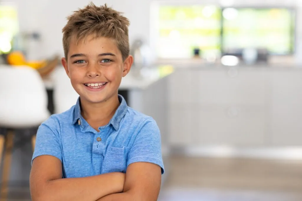 Smiling boy in a blue shirt, showcasing confidence, in a modern kitchen setting, representing orthodontic treatment options for kids, including Invisalign and braces.