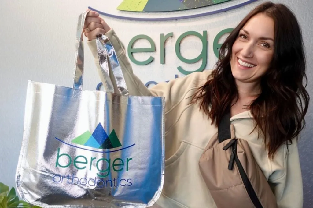 Smiling woman holding a silver tote bag with "berger orthodontics" logo, in front of a branded wall, promoting Berger Orthodontics services.