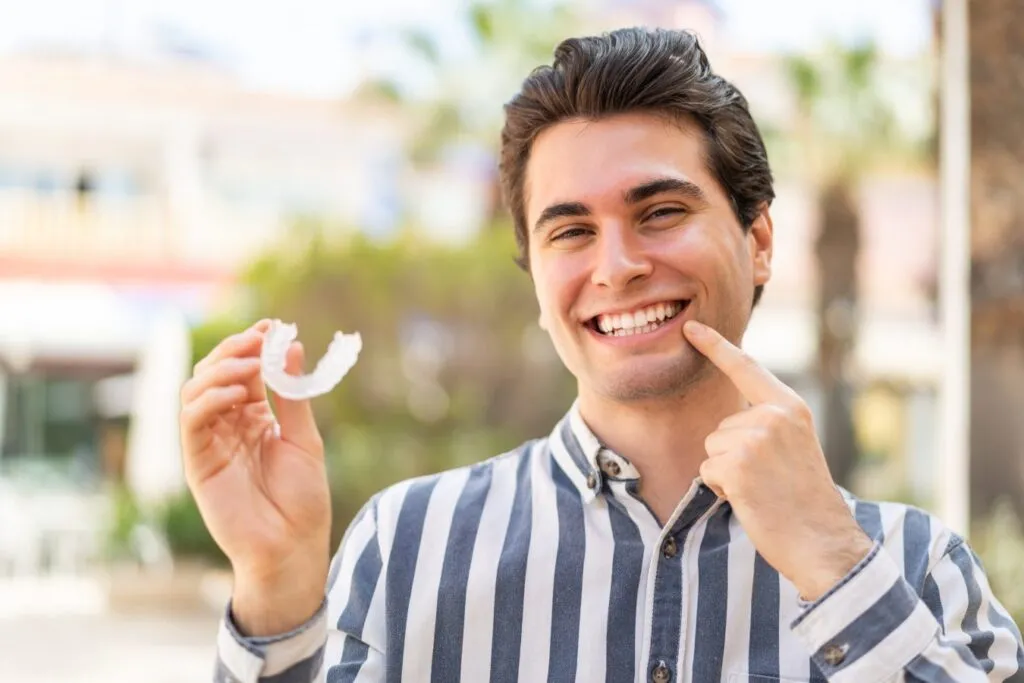 Young man smiling while holding an Invisalign clear aligner, demonstrating orthodontic treatment options for adults.