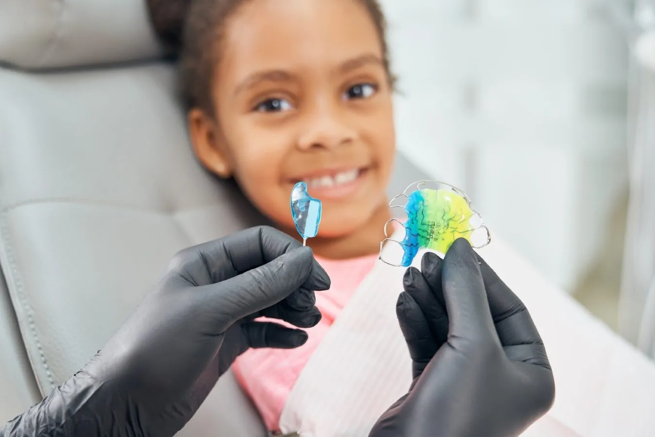 Child smiling in dental chair holding colorful orthodontic retainers, demonstrating importance of retainer care and orthodontic treatment.