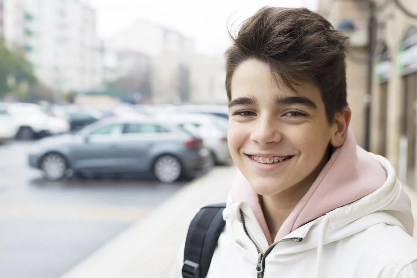Smiling teenager with braces wearing a white jacket and pink hoodie, standing outdoors near parked cars, representing the positive impact of orthodontic treatments on youth dental health.